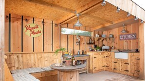Une cuisine extérieure rustique en bois, avec une table ronde, un coin salon et un panneau indiquant : « Playson's, veuillez patienter ».