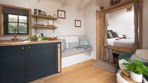 Une cuisine chaleureuse avec un plan de travail en bois, un évier et une fenêtre. On y trouve un lit avec une tête de lit en bois et une fenêtre, ainsi qu’un petit coin salon avec un banc et des coussins.