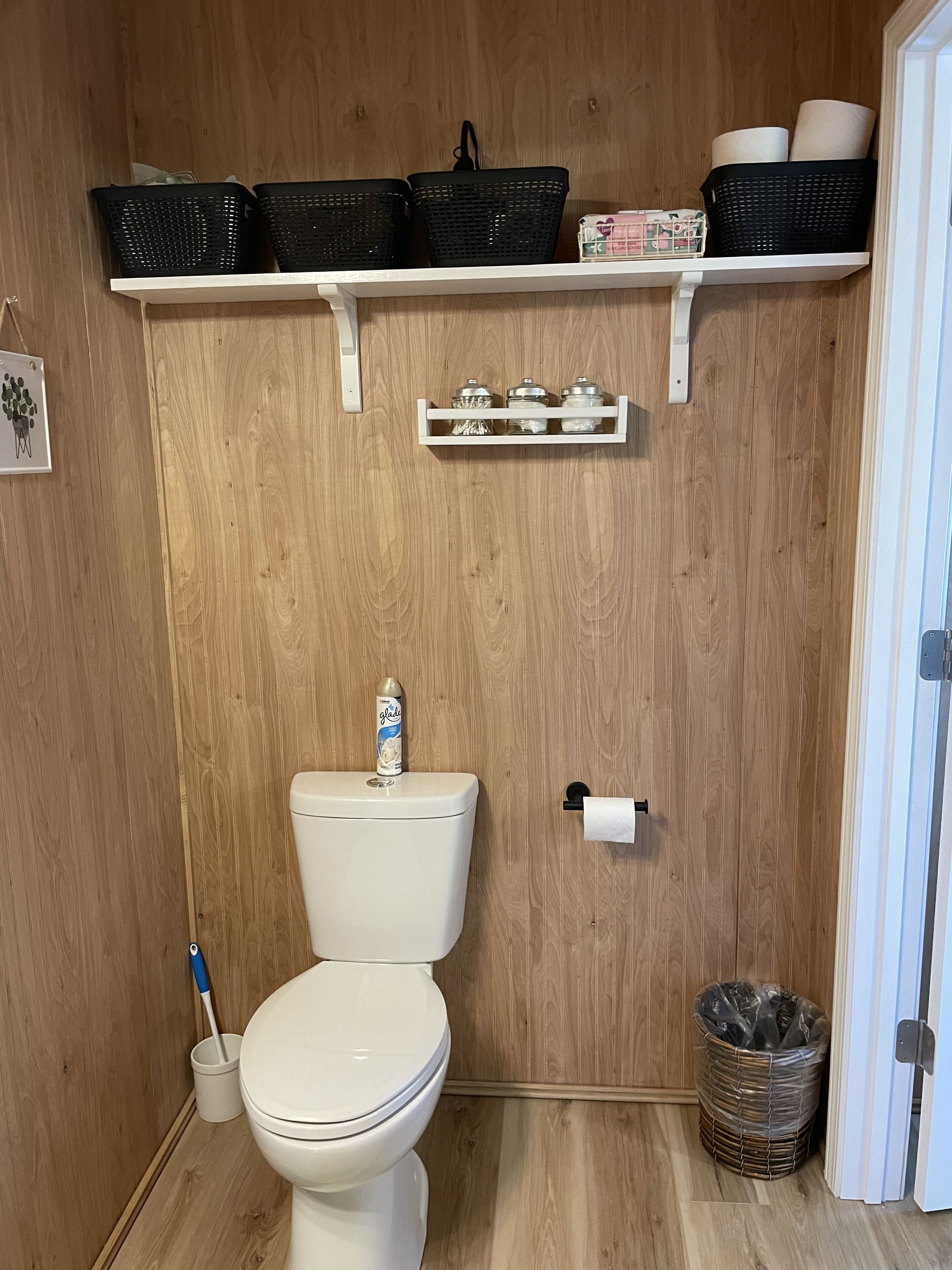 A small, wooden-paneled bathroom with a toilet, a shelf above holding baskets and toiletry items, and a waste bin.