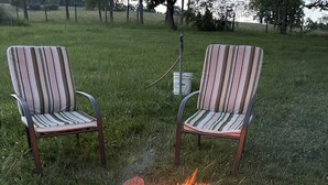 A fire pit with rocks and burning wood surrounded by grass, with two striped chairs nearby. - Meadow Farm-View Getaway (Sparta)