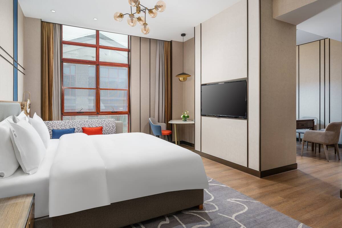 A modern hotel room with a large bed, a desk with a flat-screen TV, and a seating area with chairs.