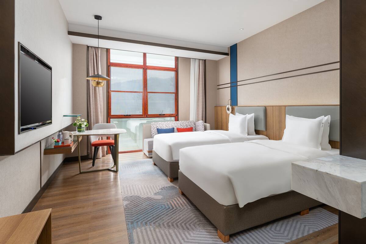 A hotel room with two beds, a TV, a desk with a chair, a window with curtains, and a bathroom with a mirror.