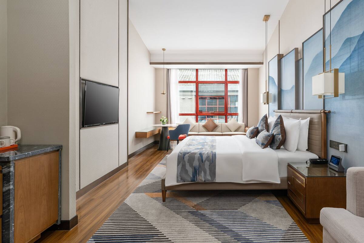 A modern hotel room with a large bed, a desk, a flat-screen TV, and a large window with red frames.