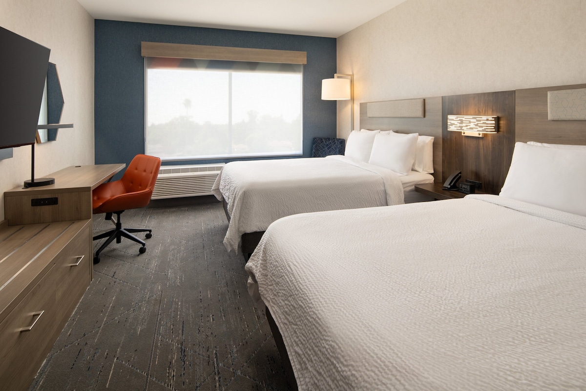 Une chambre d’hôtel avec deux lits, un bureau, une chaise et une grande fenêtre.