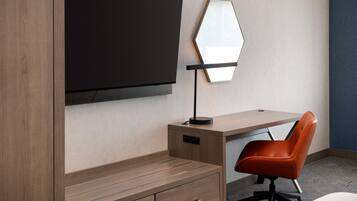 Una habitación de hotel moderna con un escritorio de madera, un televisor negro montado en la pared, un espejo hexagonal único y una silla de oficina naranja.