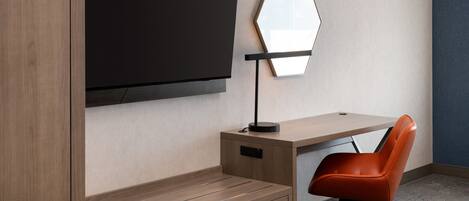 A modern hotel room with a wooden desk, a black TV mounted on the wall, a unique hexagonal mirror, and an orange office chair.