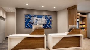 A reception area with two white reception desks, a blue and white abstract wall art, and a wooden cabinet with snacks. - Holiday Inn Express & Suites Fresno Airport by IHG (Fresno)