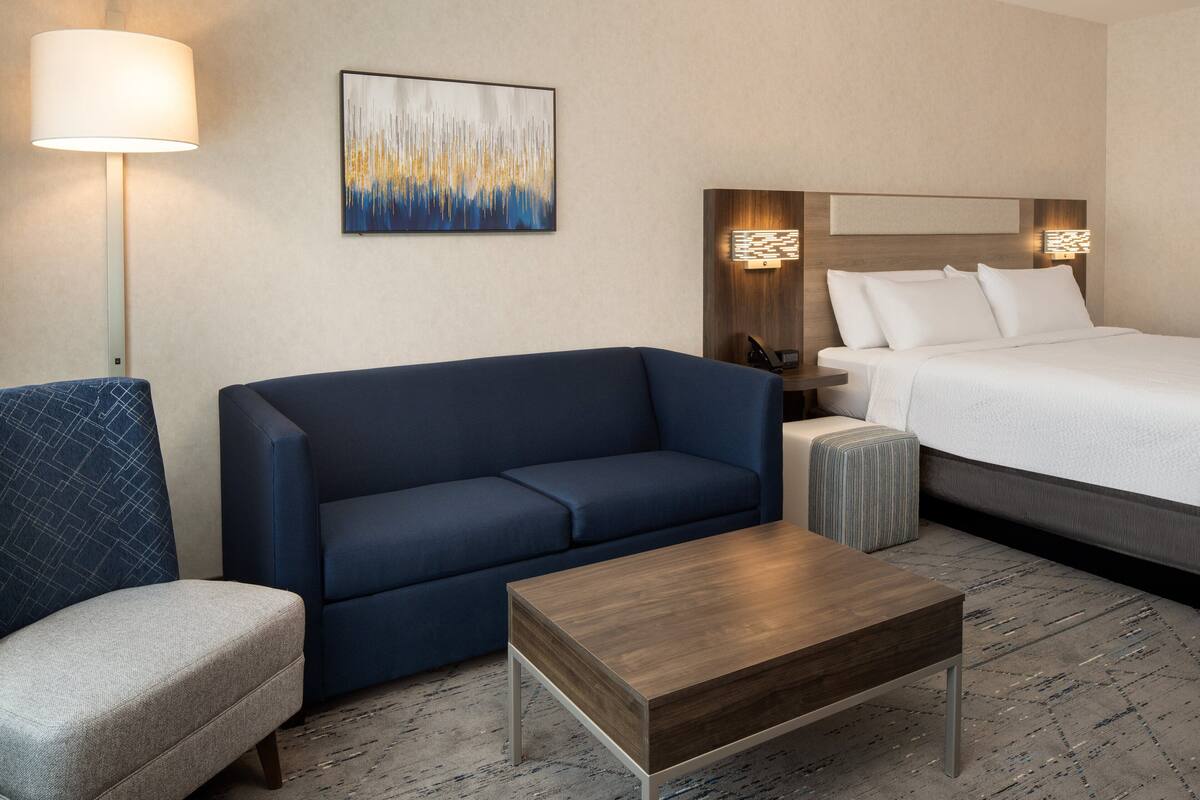 A hotel room with a bed, a blue sofa, a small armchair, a wooden coffee table, and a floor lamp.