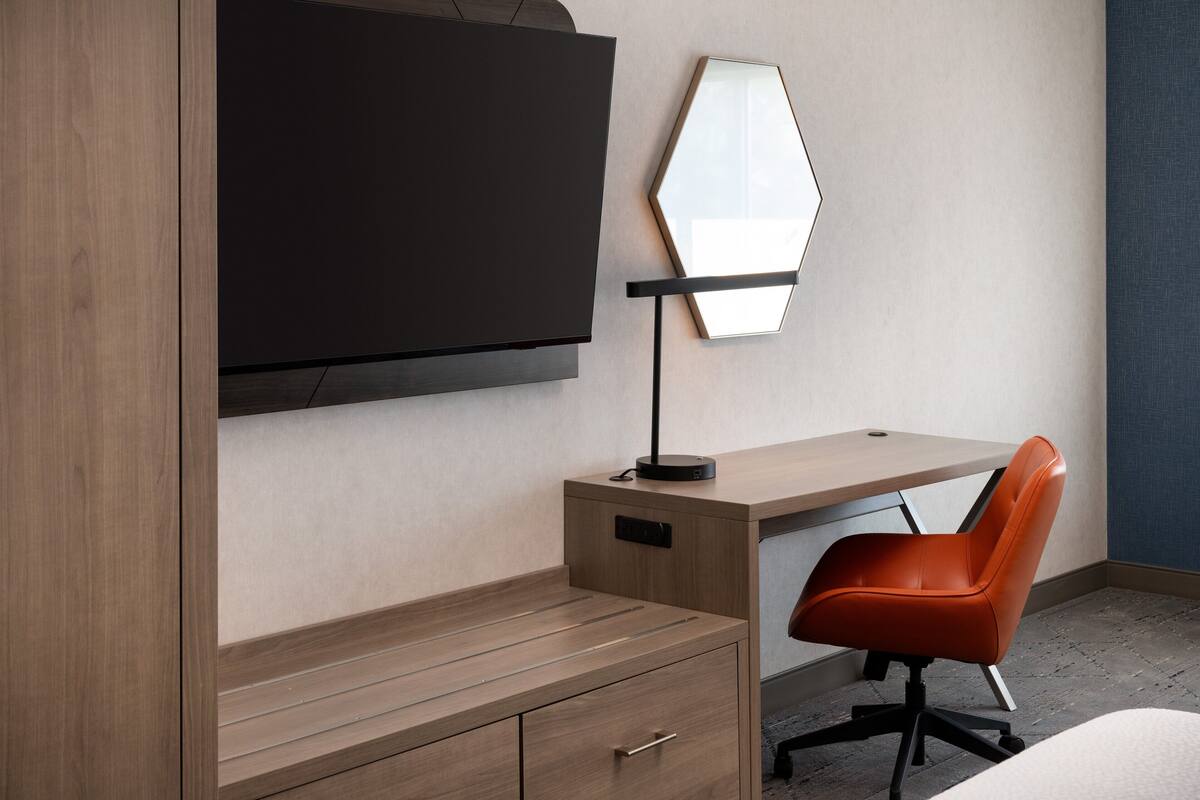 A modern hotel room with a wooden desk, a black TV mounted on the wall, a unique hexagonal mirror, and an orange office chair.