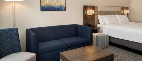 A hotel room with a bed, a blue sofa, a small armchair, a wooden coffee table, and a floor lamp.