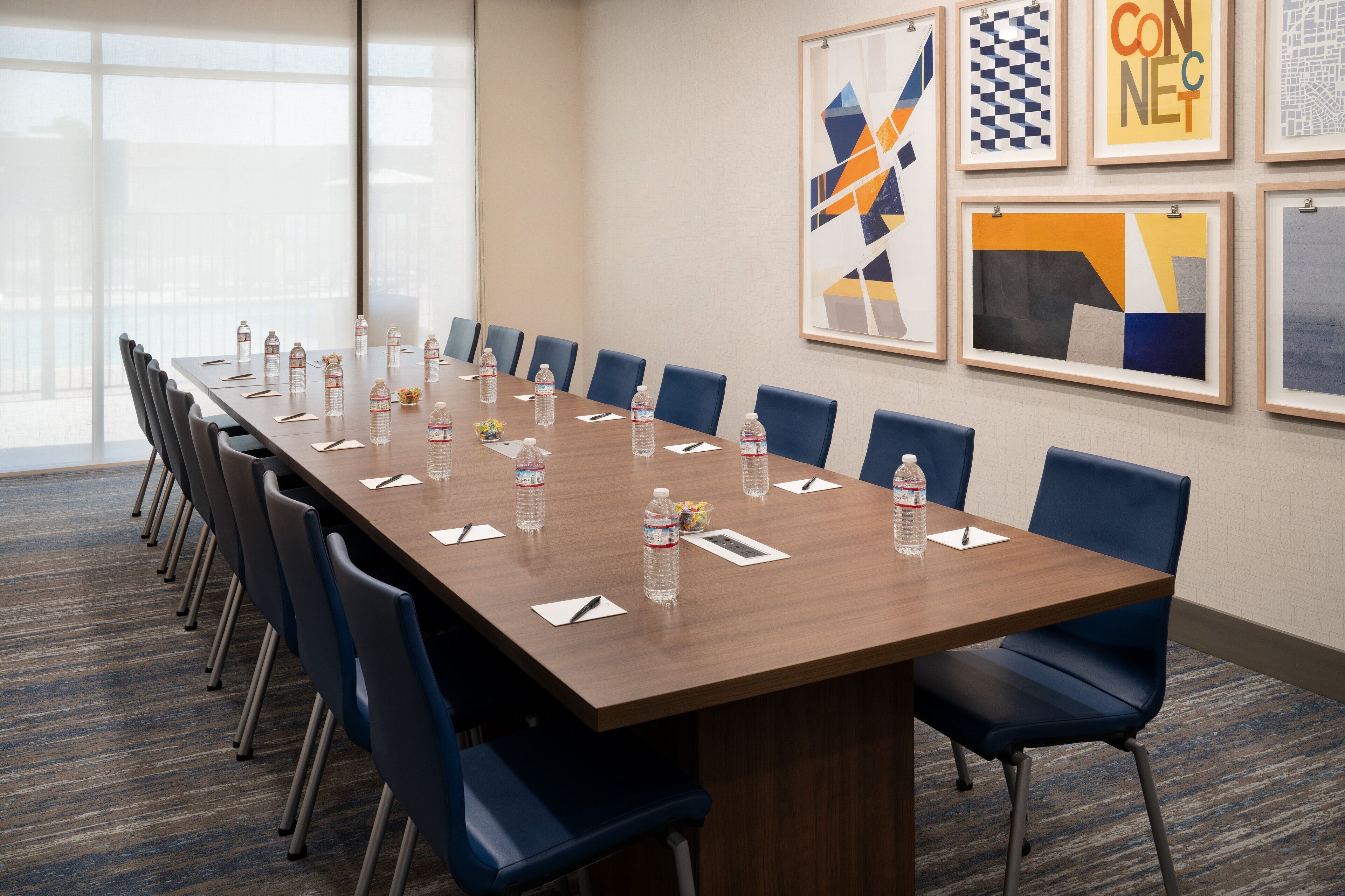 A conference room with a long table, blue chairs, water bottles, and framed artwork on the wall.