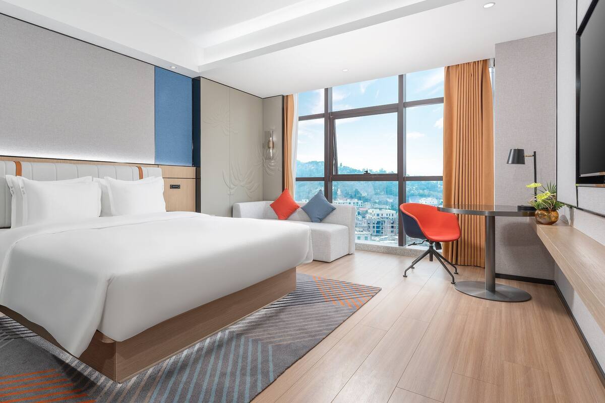 A modern hotel room with a large bed, a desk, and a view of the city.