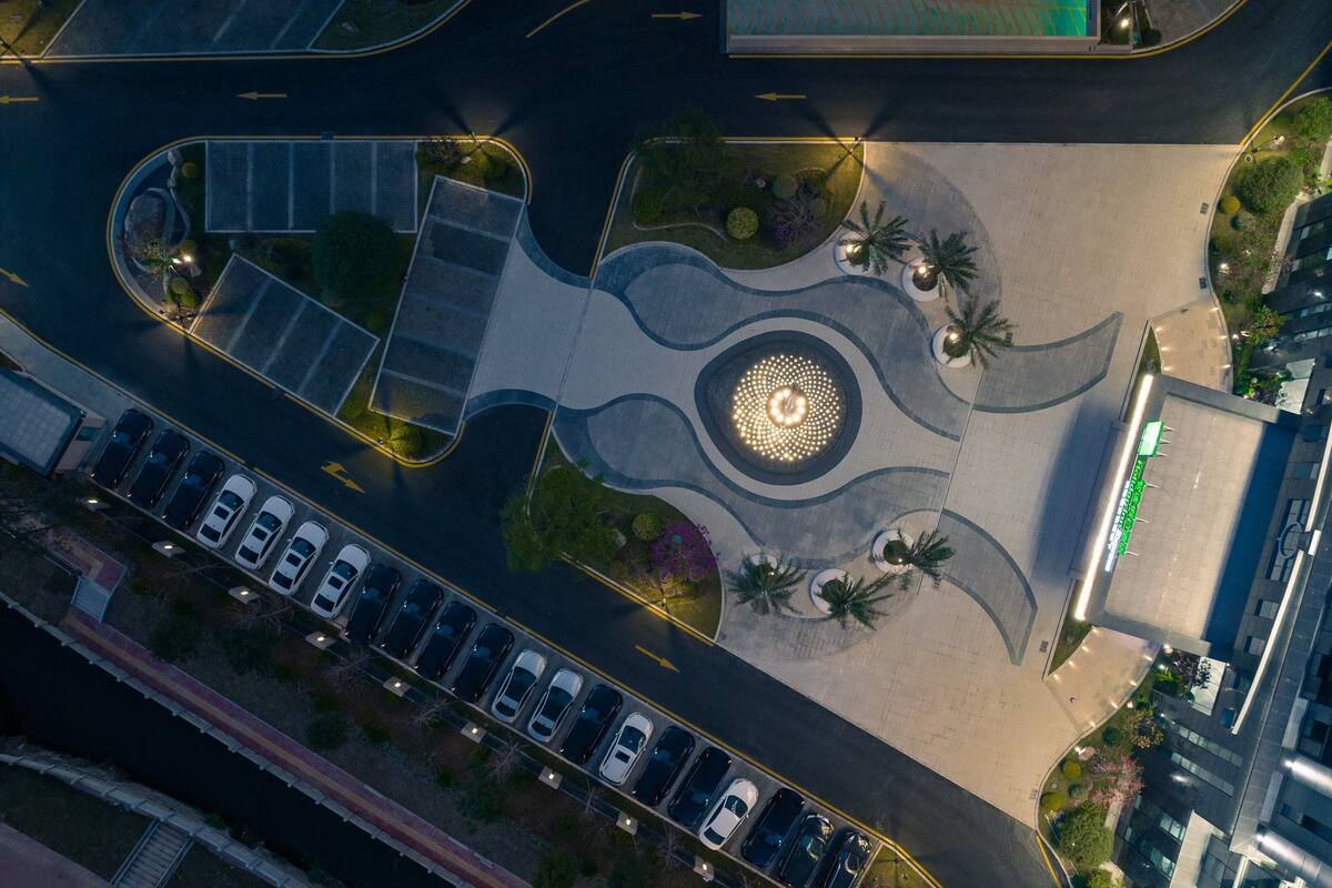 An aerial view of a parking lot with multiple rows of cars, a landscaped area with palm trees, and a circular light feature.