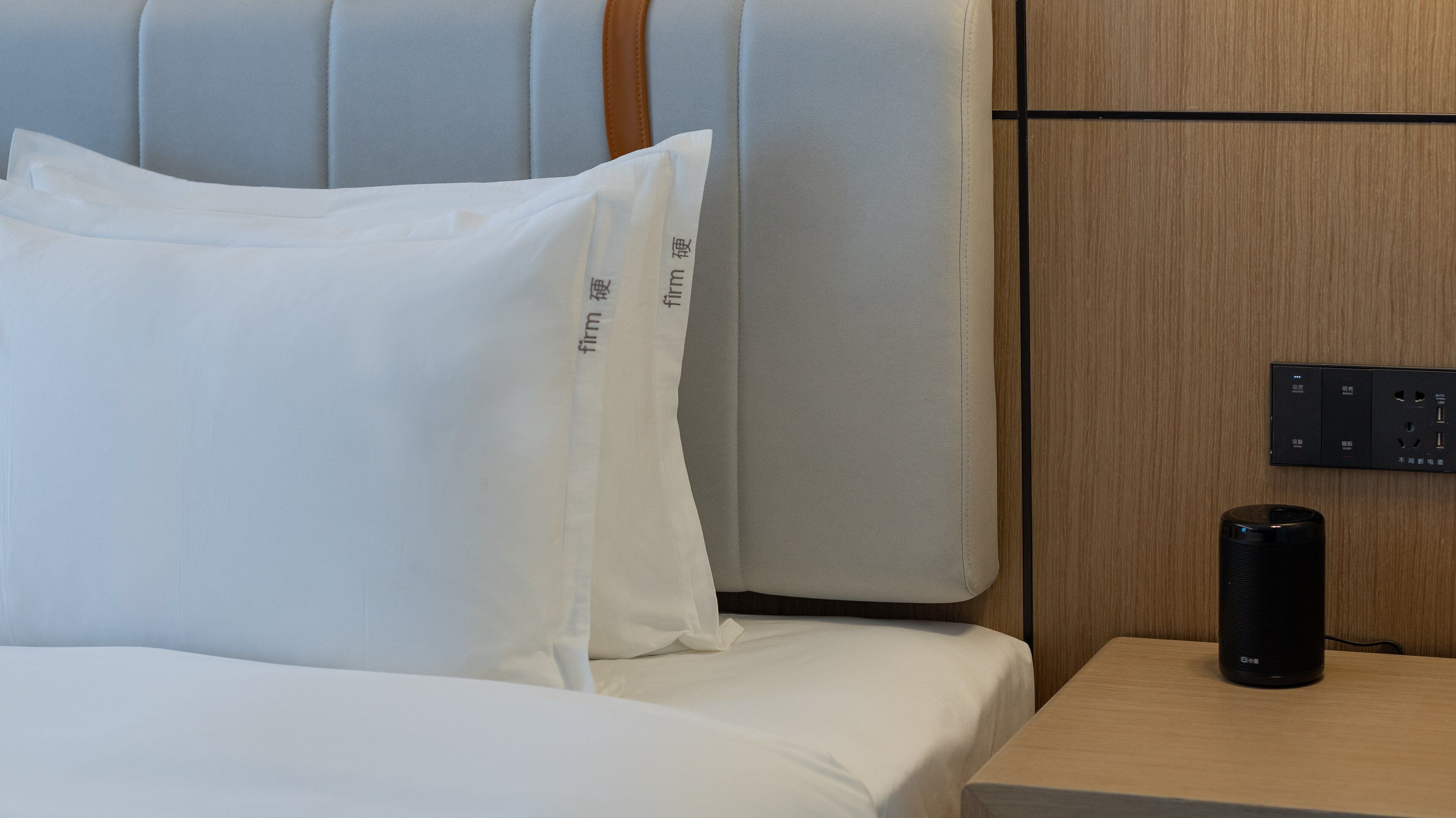 A neatly made bed with white linens, a bedside table with a black speaker, and a wall-mounted control panel.