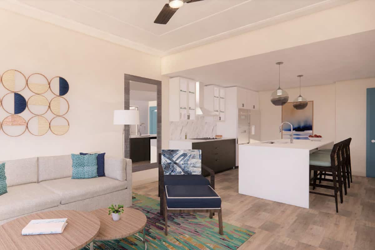 A modern living room with a sofa, coffee table, and a colorful rug. There is a kitchen area with cabinets and a dining table.