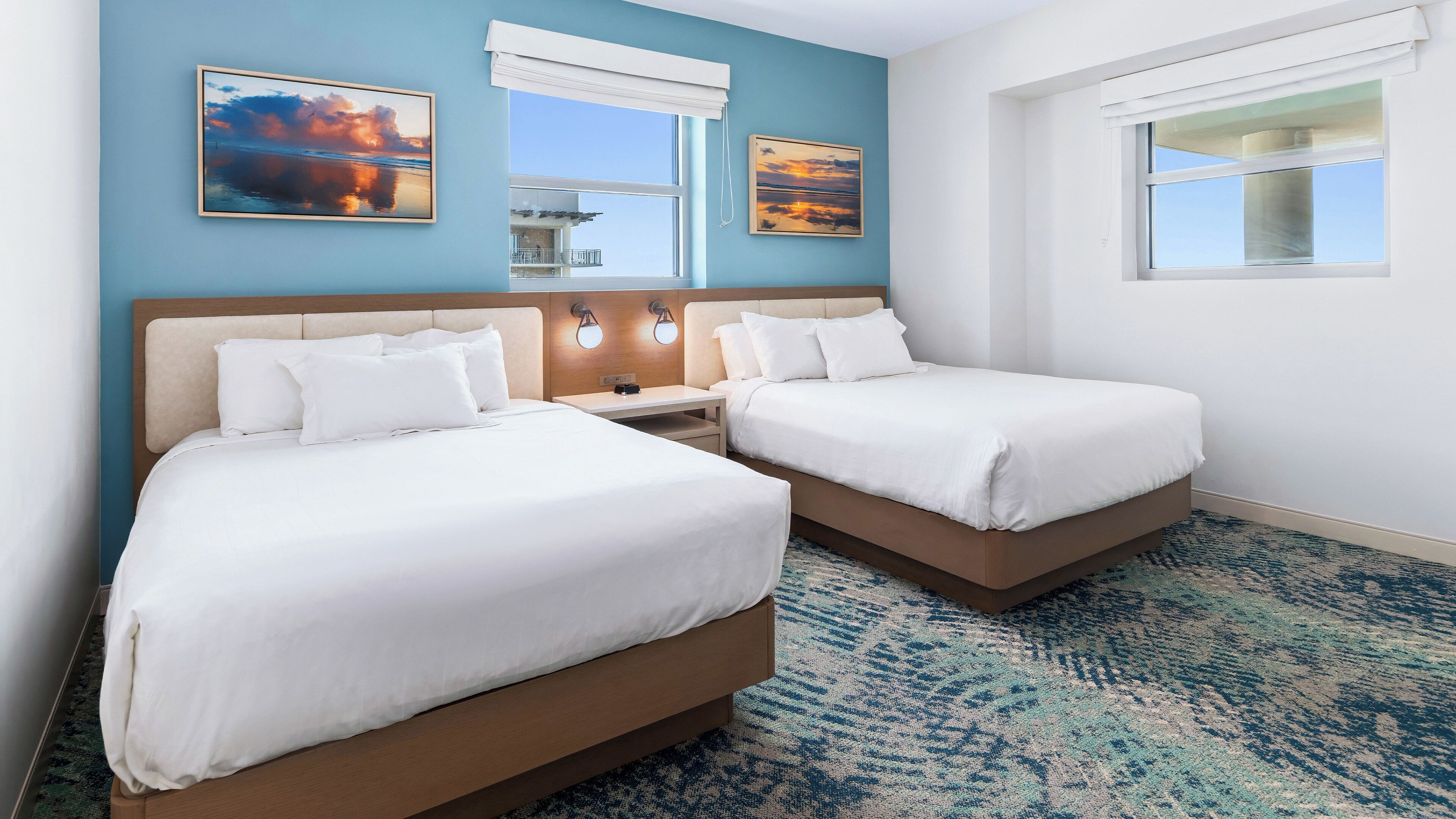 A hotel room with two beds, a nightstand, wall-mounted lights, and a large window with a view.