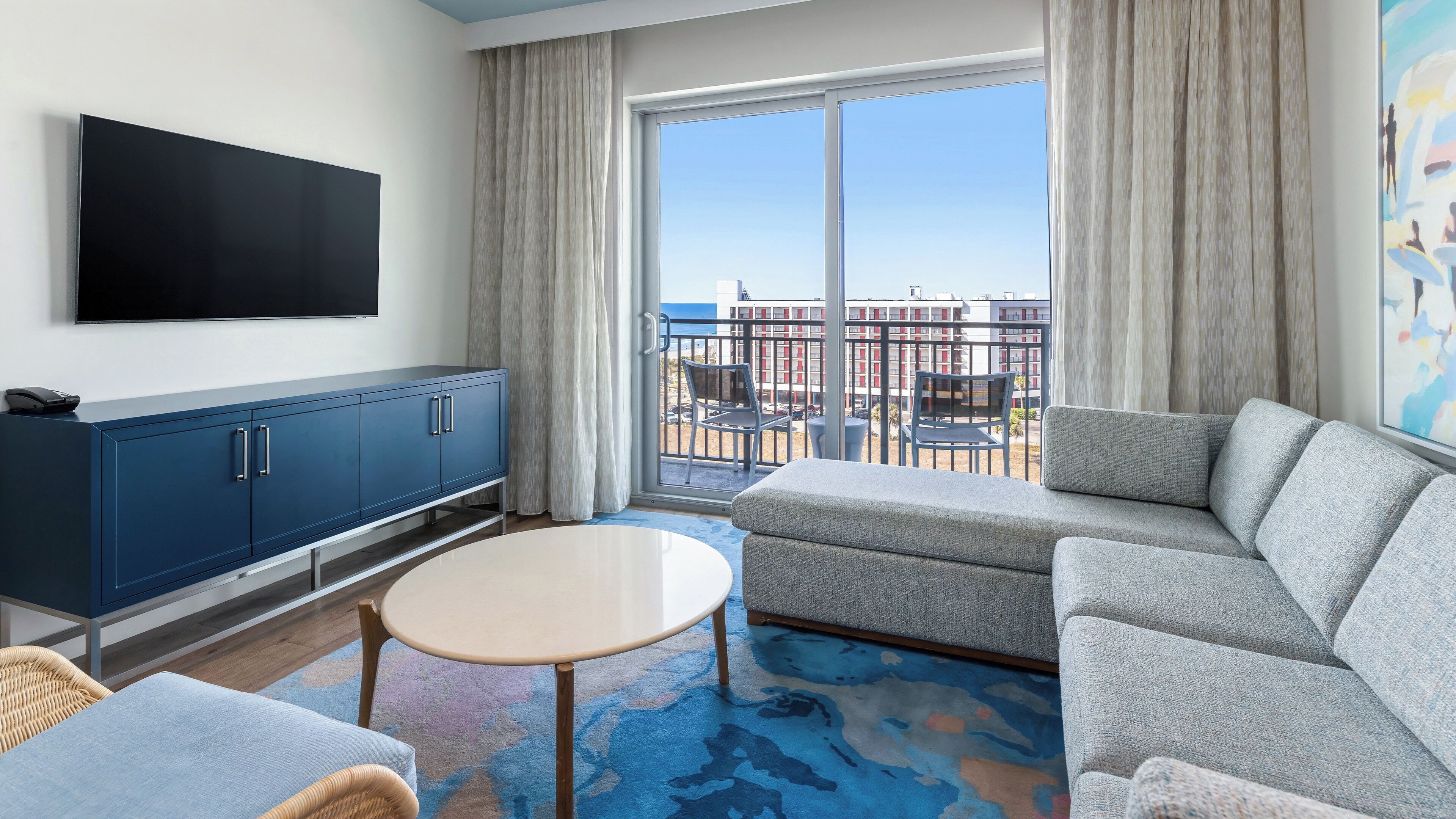 Ein modernes Hotelzimmer mit großem Fenster, Flachbildfernseher, einer Couch, einem kleinen runden Tisch und einem Schrank.