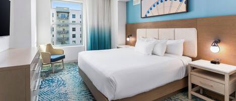 A hotel room with a bed, a desk, a chair, a nightstand, a wall-mounted lamp, and a window with a view of buildings.