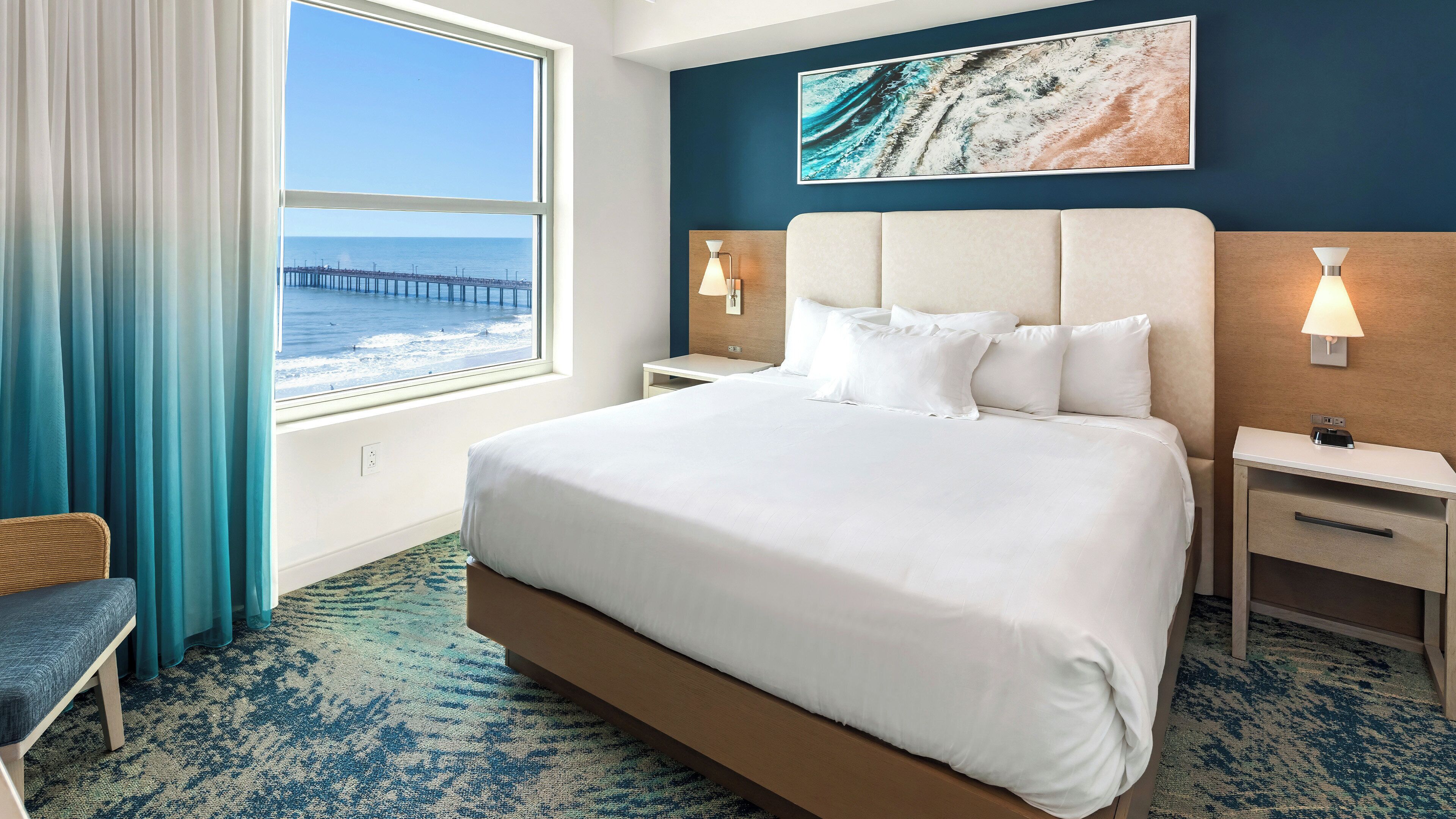 A hotel room with a large bed, a view of the ocean, a wooden headboard, and a painting on the wall.