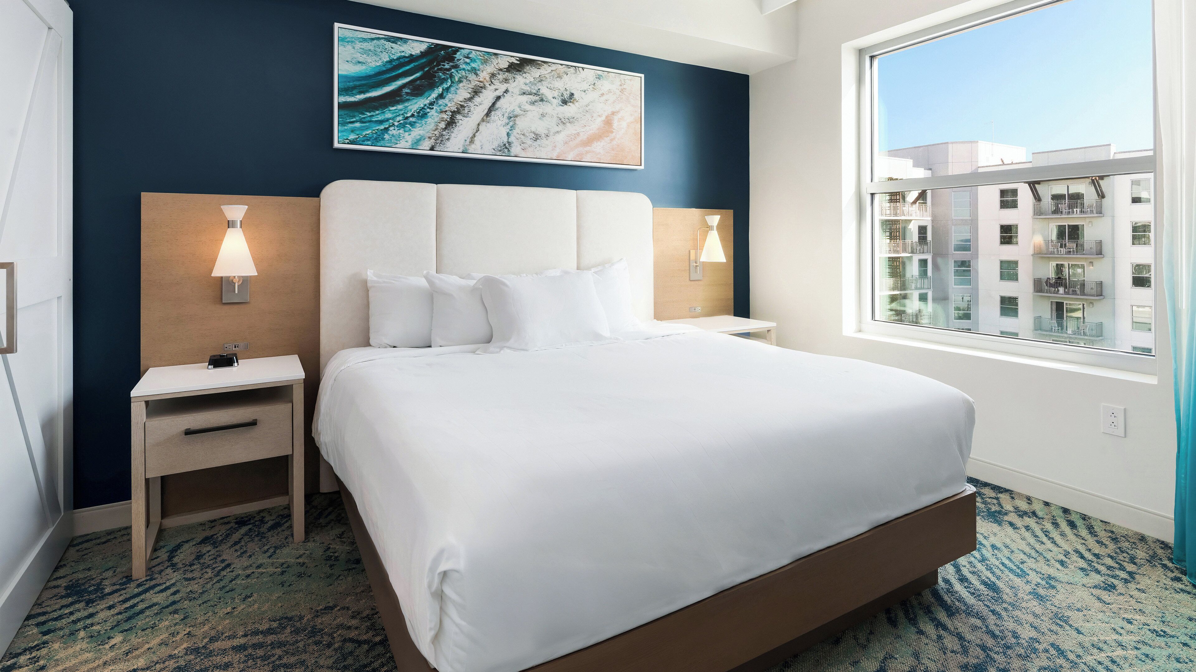 Habitación de hotel con una cama grande, dos mesitas de noche, una lámpara de pared, un ventanal con vista a edificios y un cuadro sobre la cama.
