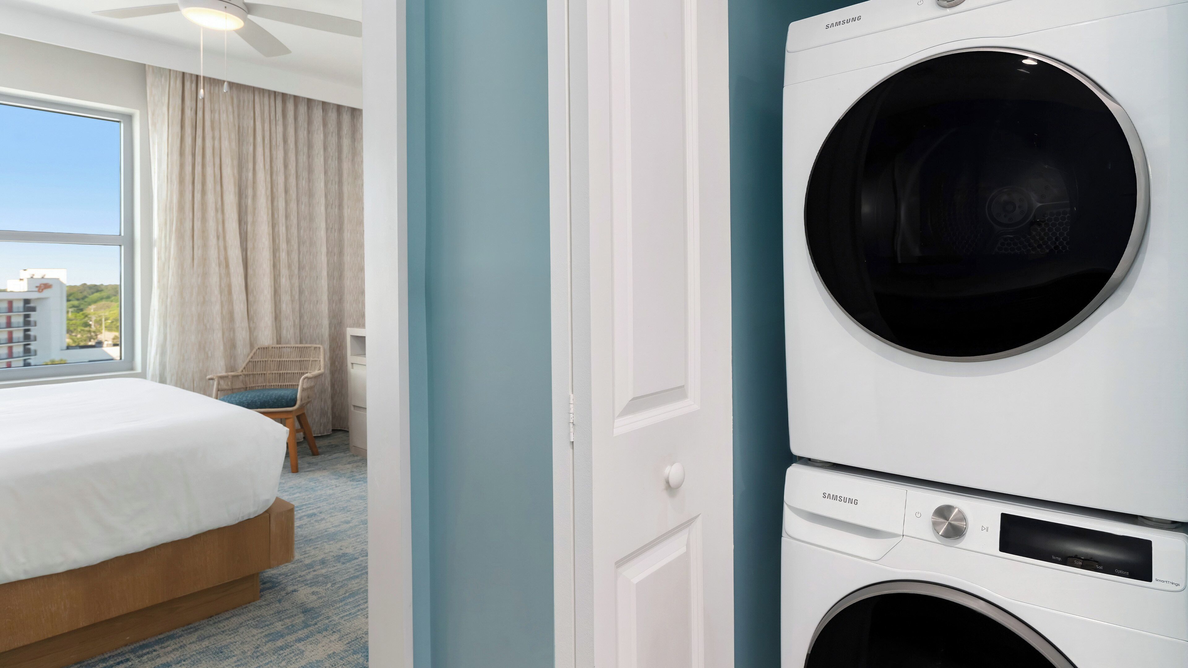 A hotel room with a bed, a chair, a washing machine, and a view of a building through a window.