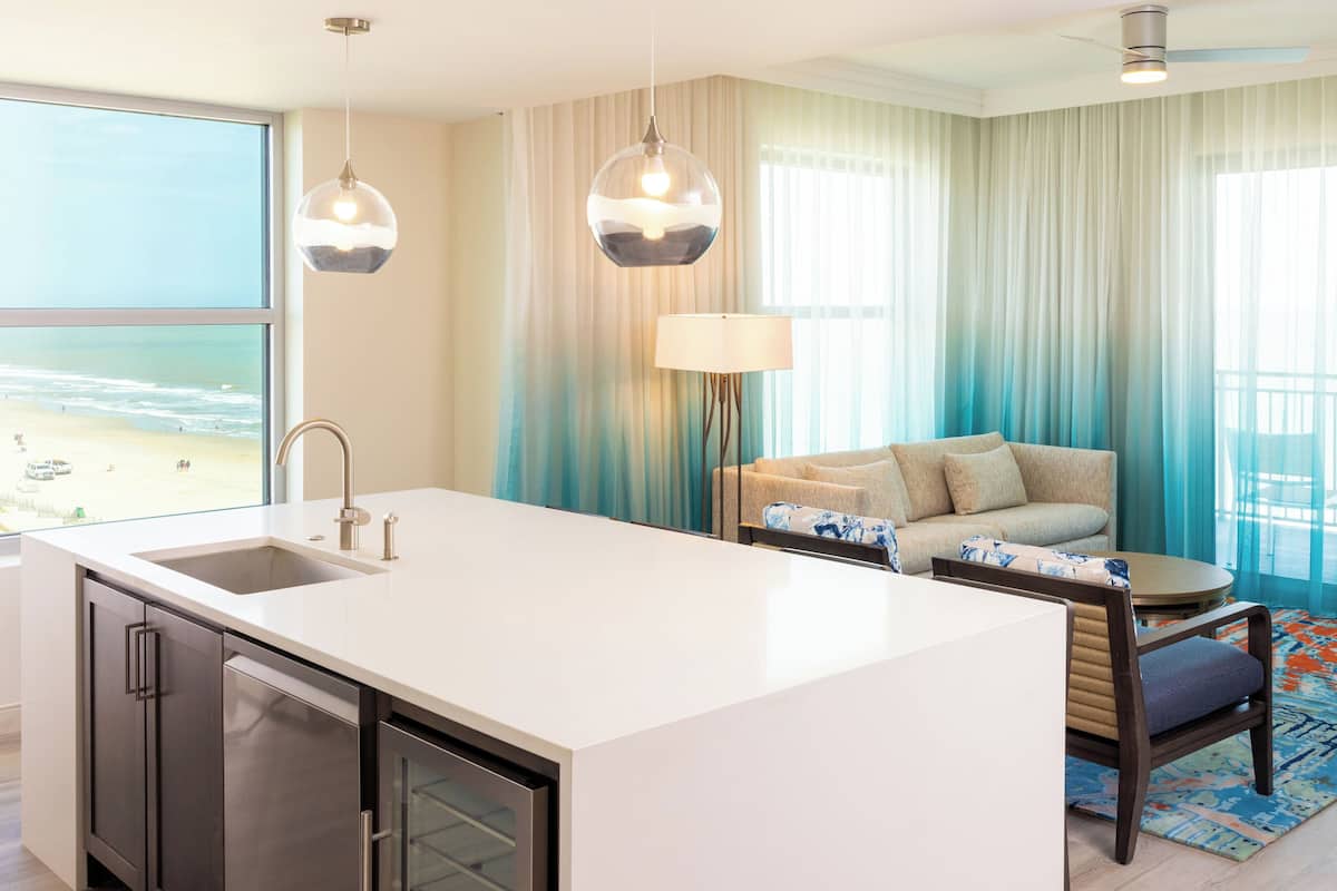 A modern kitchen with a large island, a built-in sink, and a view of the beach. There is a seating area with a sofa and a table, and a large window with sheer curtains.