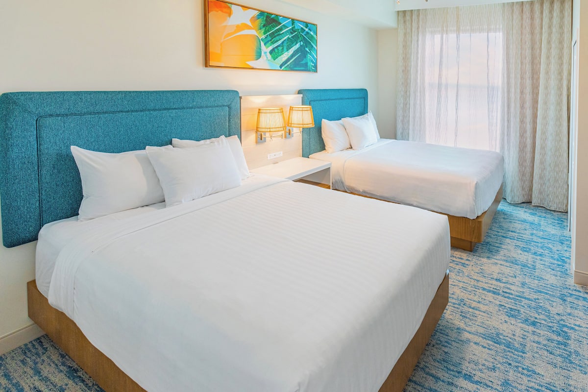 A hotel room with two beds, a large window with curtains, a painting on the wall, and a wooden nightstand with lamps.