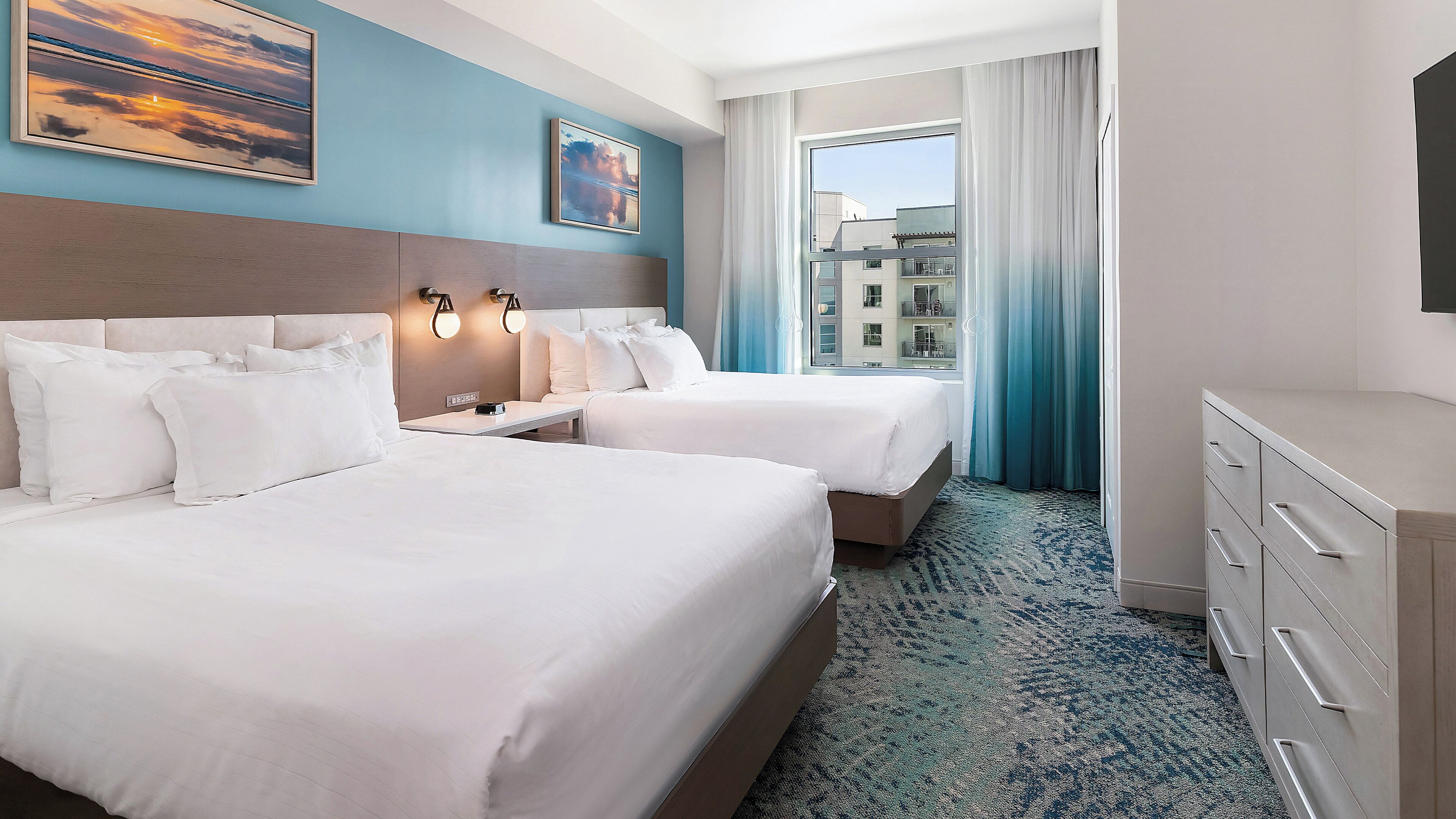 A hotel room with two beds, a desk, a dresser, and a large window with curtains.