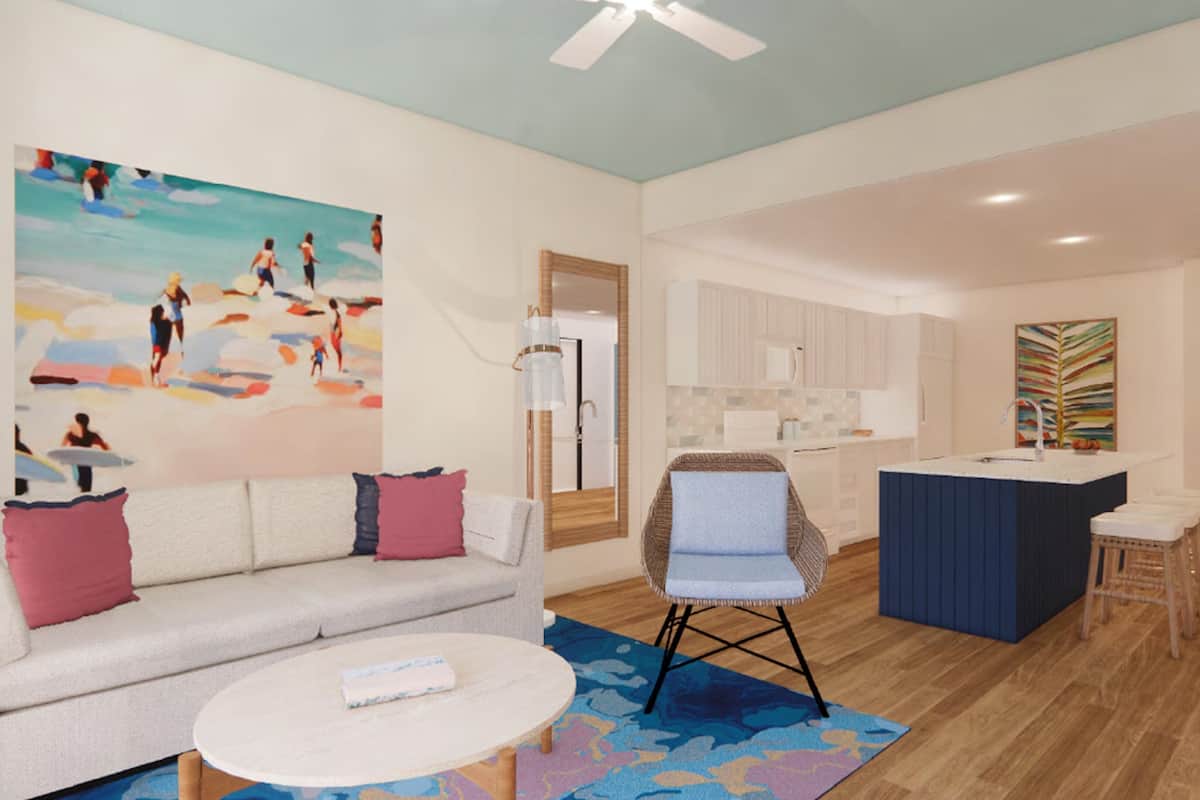 A modern living room with a sofa, coffee table, and a painting of beachgoers. A kitchen area with a blue island and stools is visible in the background.