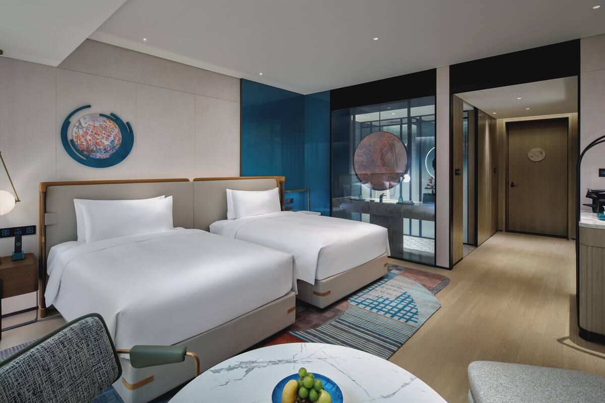 A hotel room with two beds, a desk, a chair, a round table with a fruit bowl, a large abstract painting, and a view of the city through a glass wall.
