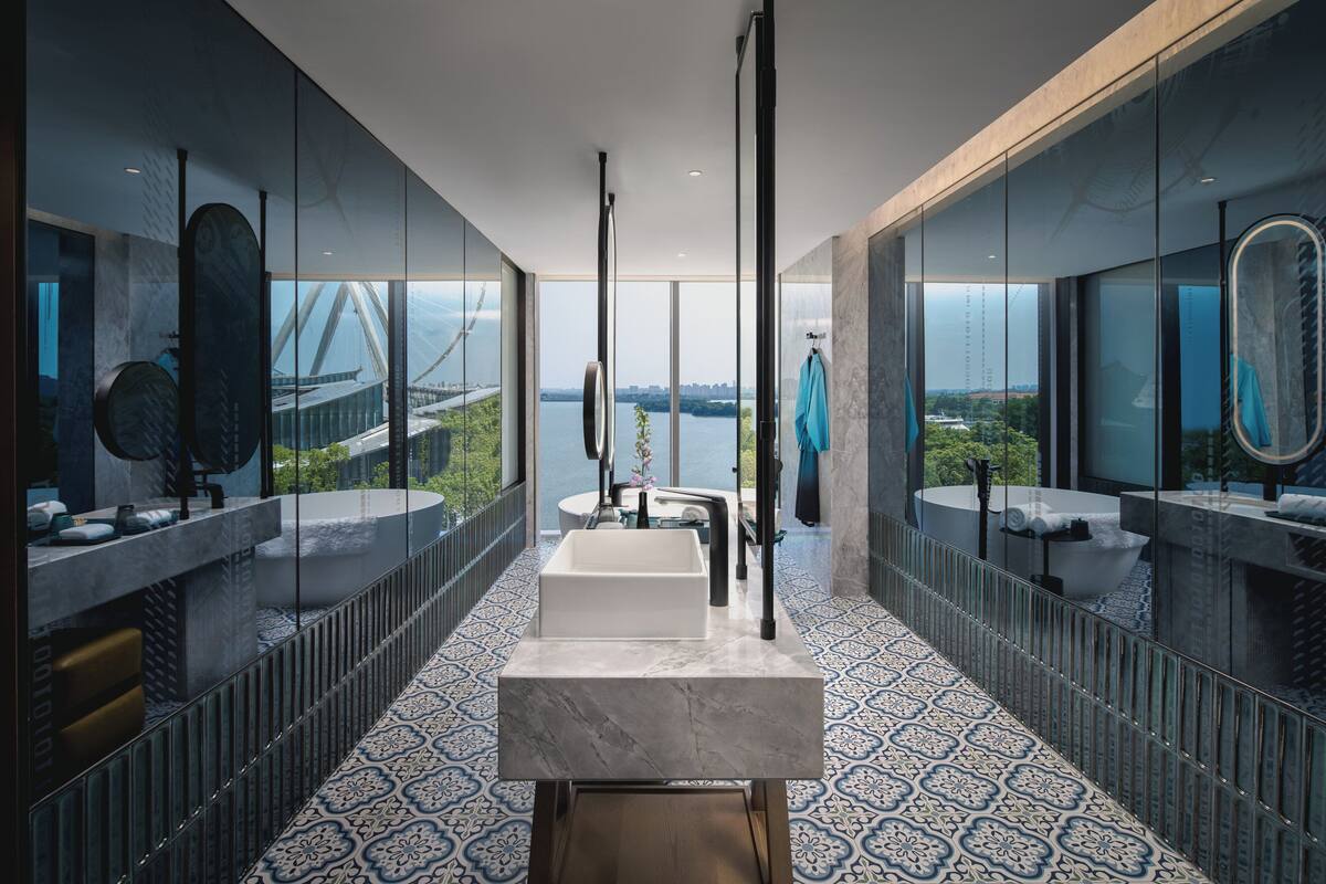 A modern bathroom with a large bathtub, a rectangular sink, and a mirror. The room has a patterned floor and large windows offering a view of a bridge and greenery.