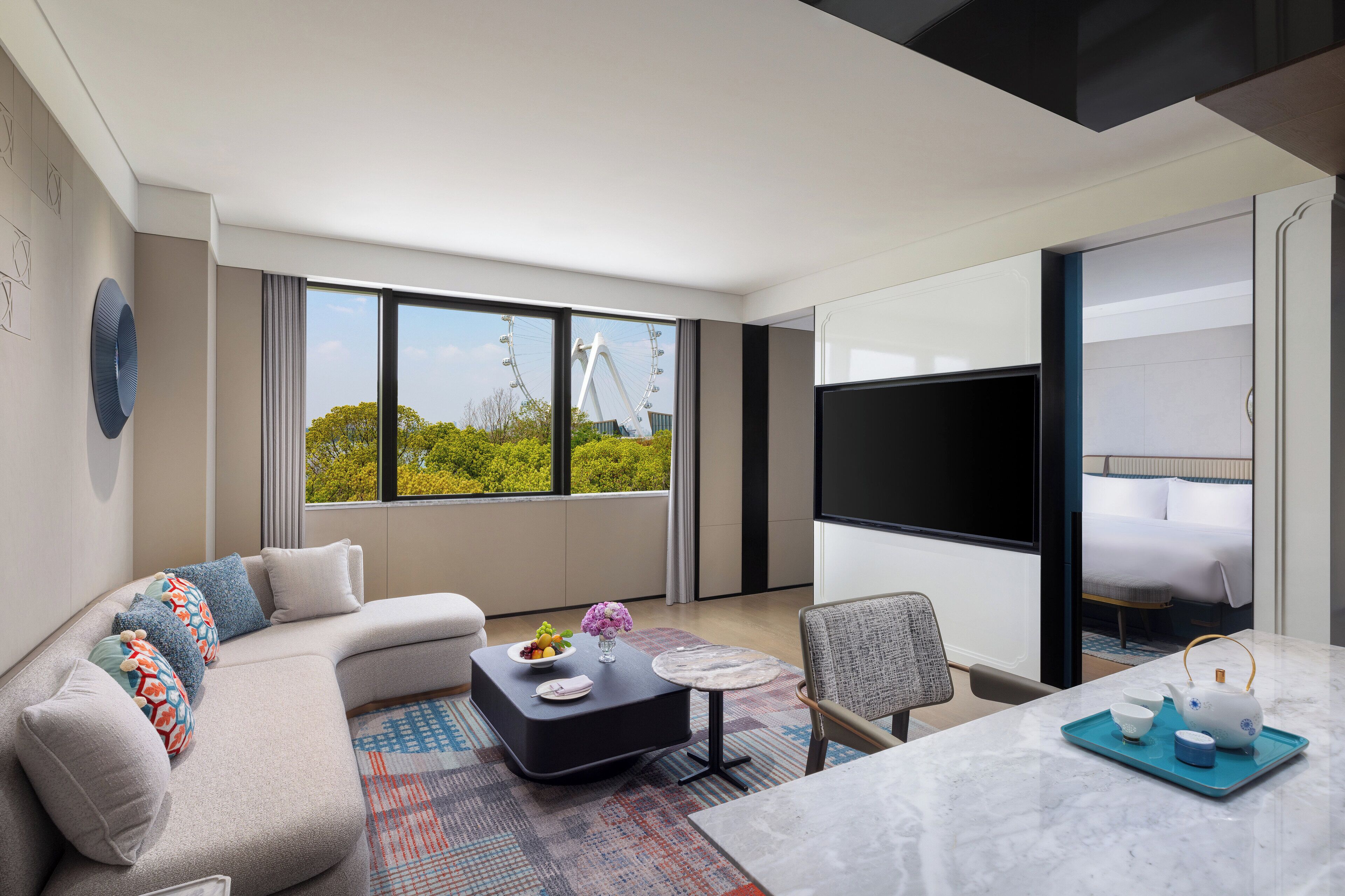 Une chambre d’hôtel moderne avec un canapé incurvé, une petite table ronde, une chaise, un lit avec un téléviseur et de grandes fenêtres donnant sur une grande roue.