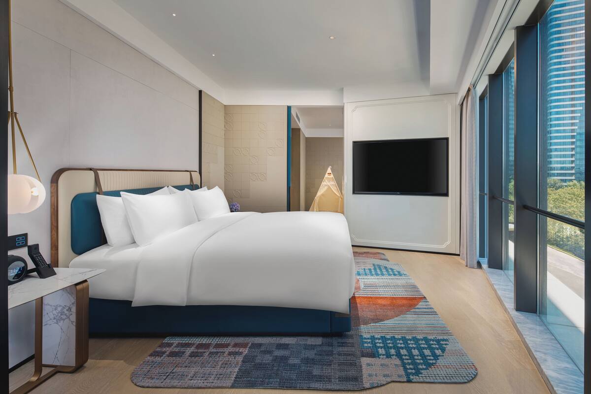 A modern hotel room with a large bed, a desk with a lamp, a flat-screen TV, and large windows offering a city view.