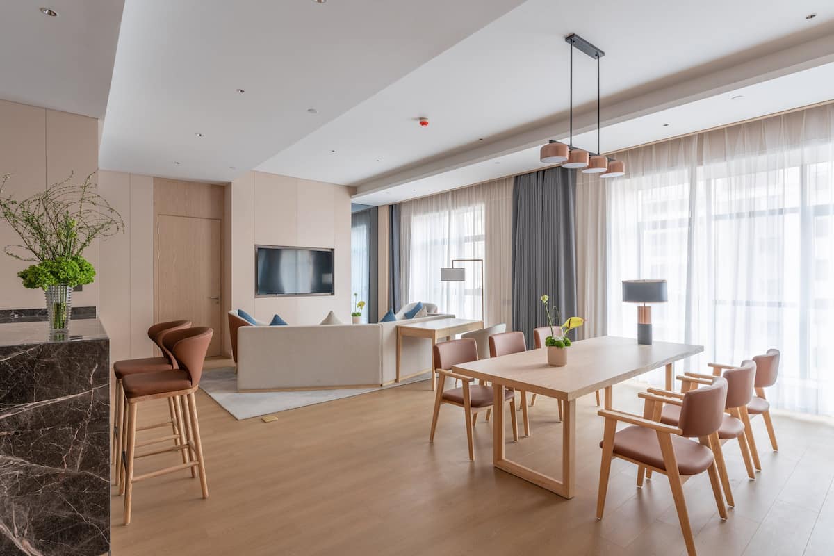 Ein moderner Essbereich mit einem langen Tisch und Stühlen, ein Küchenbereich mit einem Fernseher und große Fenster mit leichtem Vorhang.