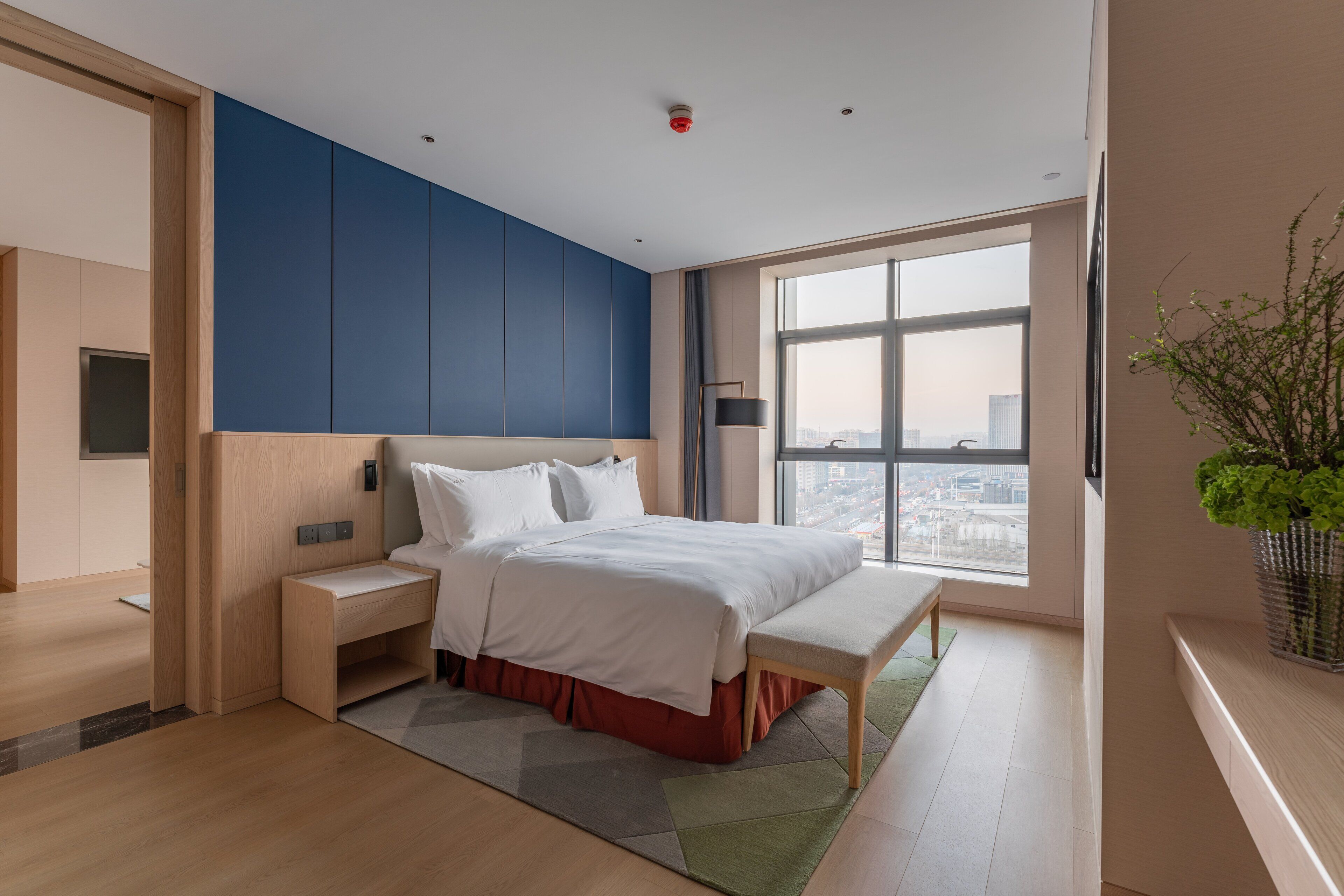 Een moderne hotelkamer met een groot bed, een bankje, een nachtkastje, uitzicht op de stad door grote ramen en een plant op de vensterbank.