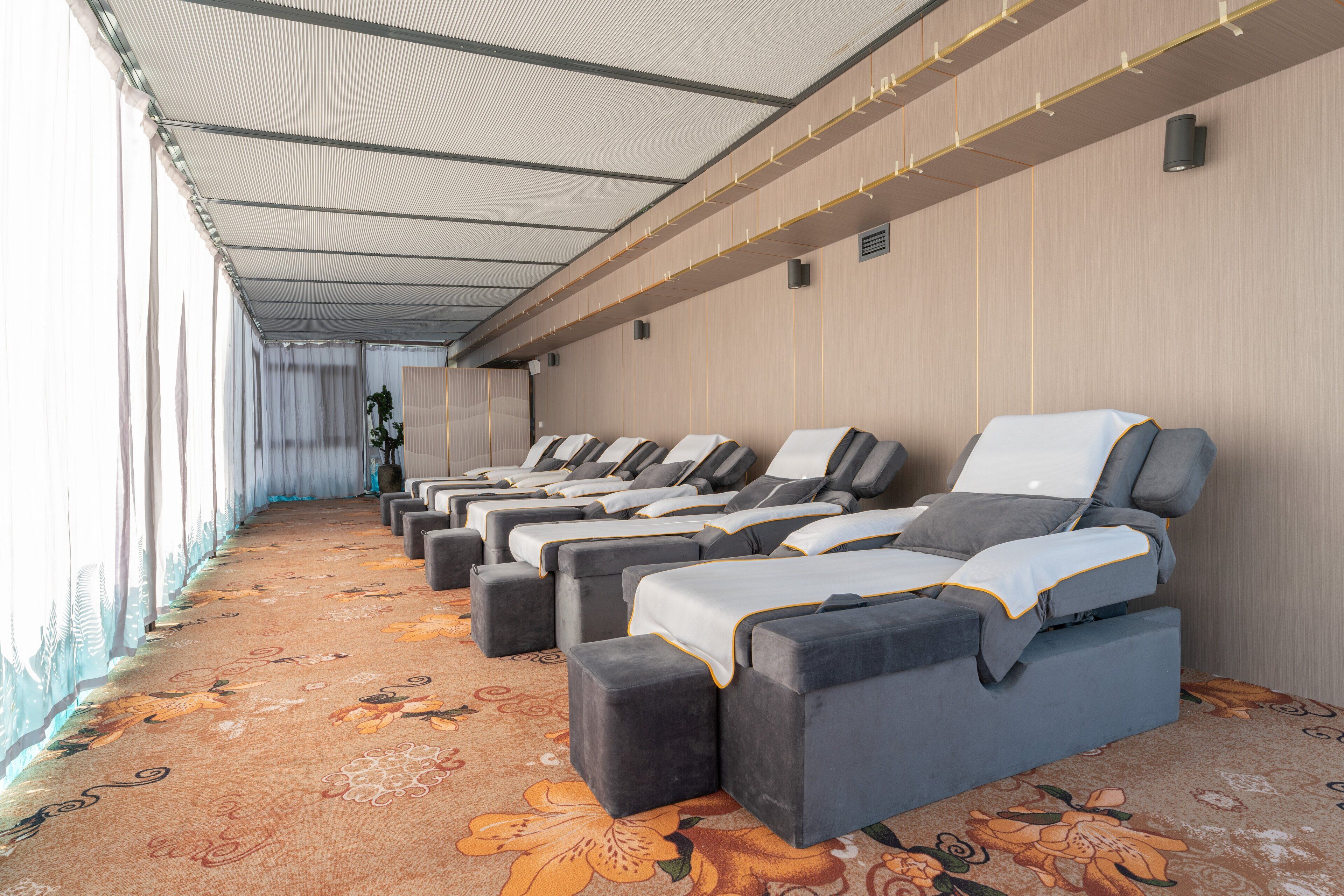 Une rangée de salles de massage, chacune équipée d’un lit individuel et d’un repose-pieds, ainsi qu’un tapis à motifs et un plafond à structure quadrillée.