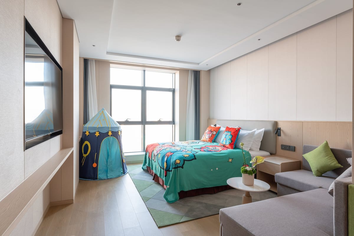 Ein modernes Hotelzimmer mit einem großen Bett, einer kleinen Couch, einem Fernseher, einem Fenster und einem Spielzelt.