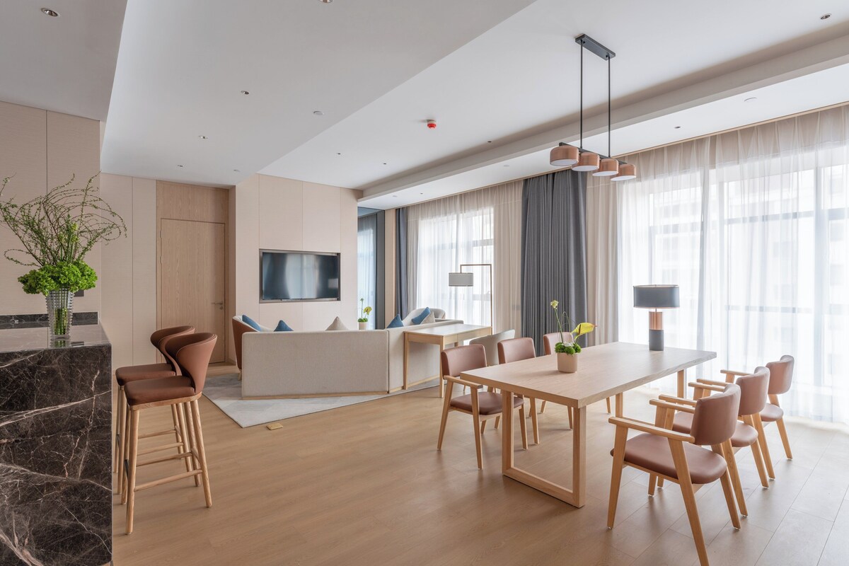 A modern dining area with a long table and chairs, a kitchen area with a TV, and large windows with sheer curtains.