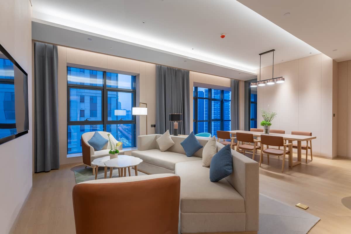 Ein modernes Hotelzimmer mit einer Couch, Sesseln, einem Esstisch und großen Fenstern mit Blick auf die Stadt.