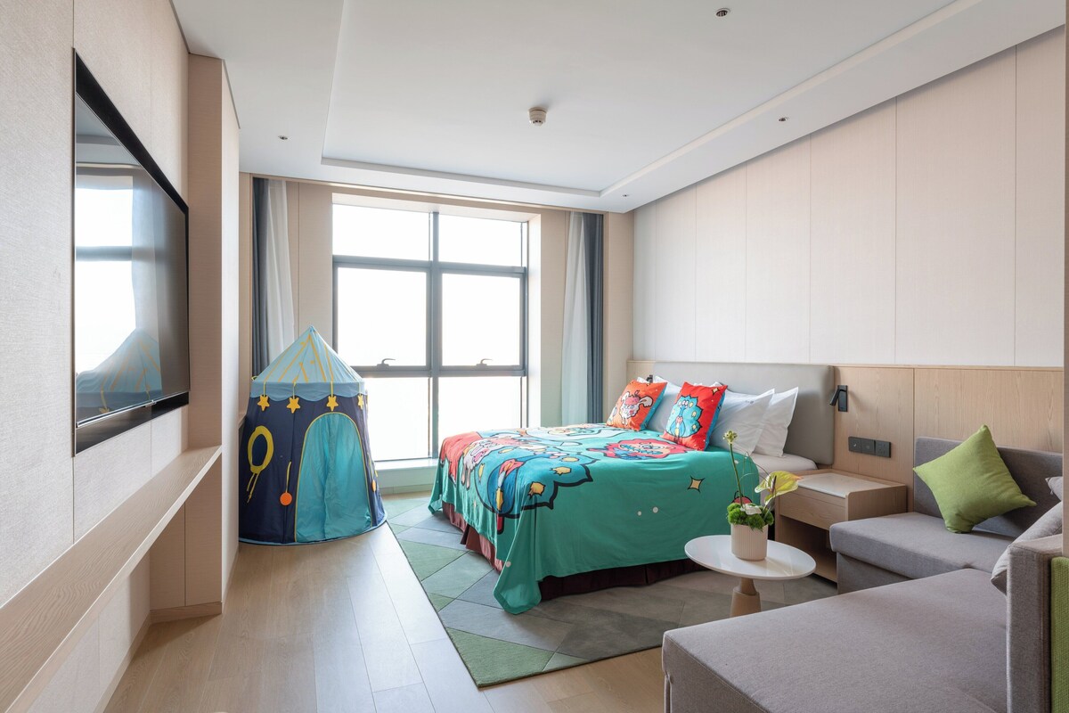 A modern hotel room with a large bed, a small sofa, a TV, a window, and a play tent.