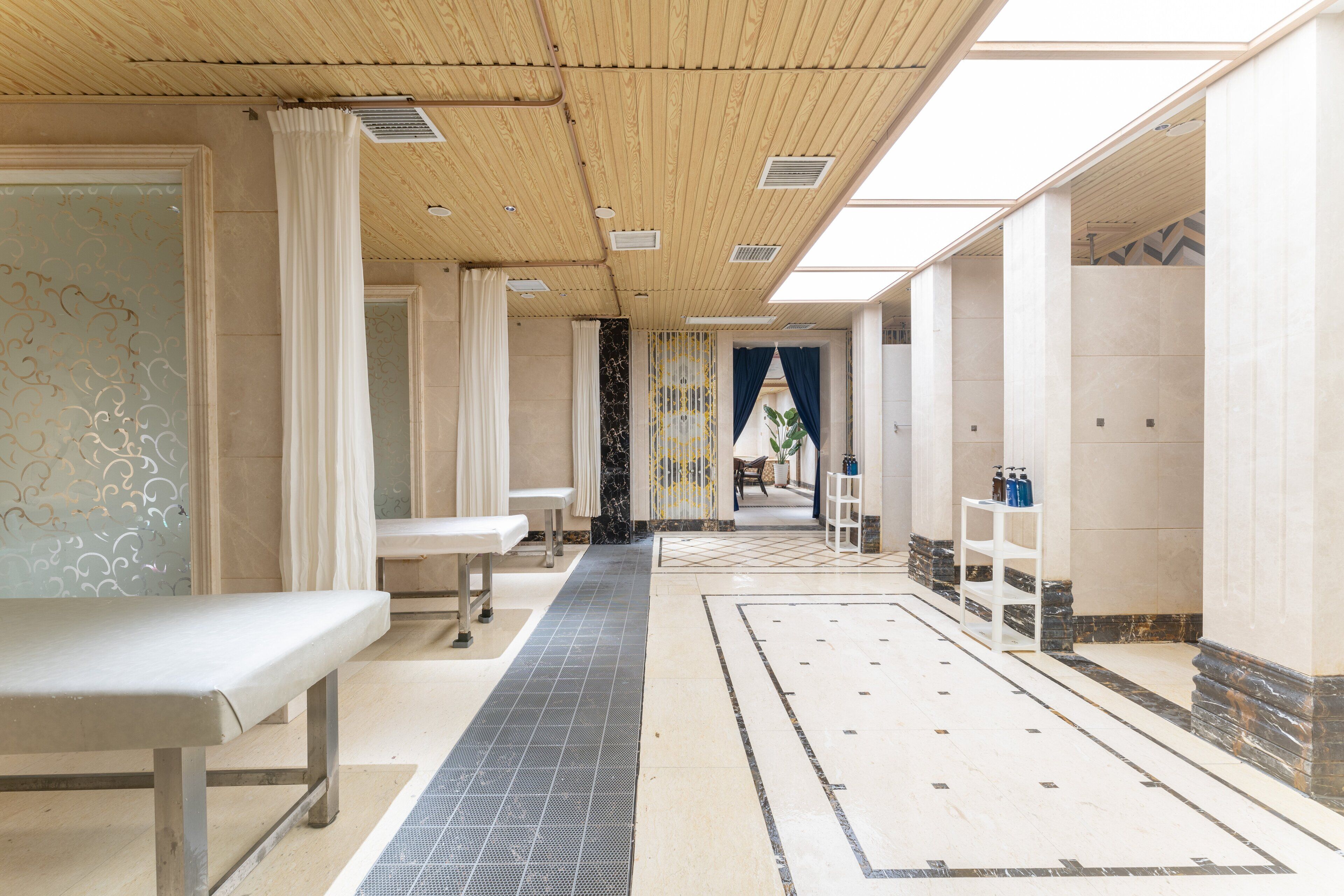 A hotel corridor with a long bench, a tiled floor, and a view of a room with a bed and a window.