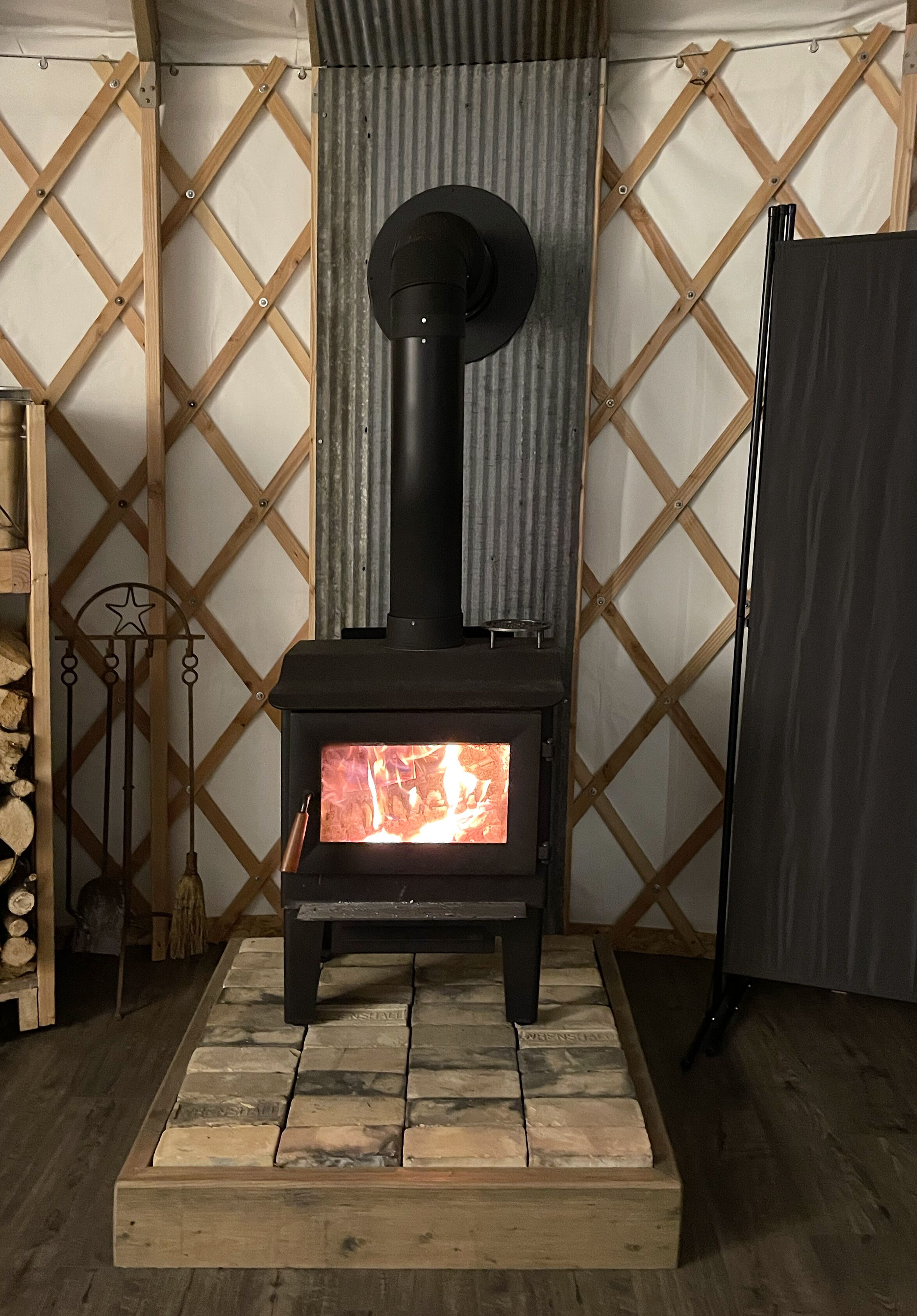 A wood-burning stove with a visible fire, set on a stone platform, against a lattice wall with a corrugated metal backdrop.