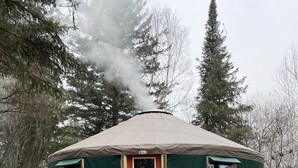Une yourte avec un escalier en bois menant à une porte rouge, un barbecue et une bouteille de propane à l’extérieur.