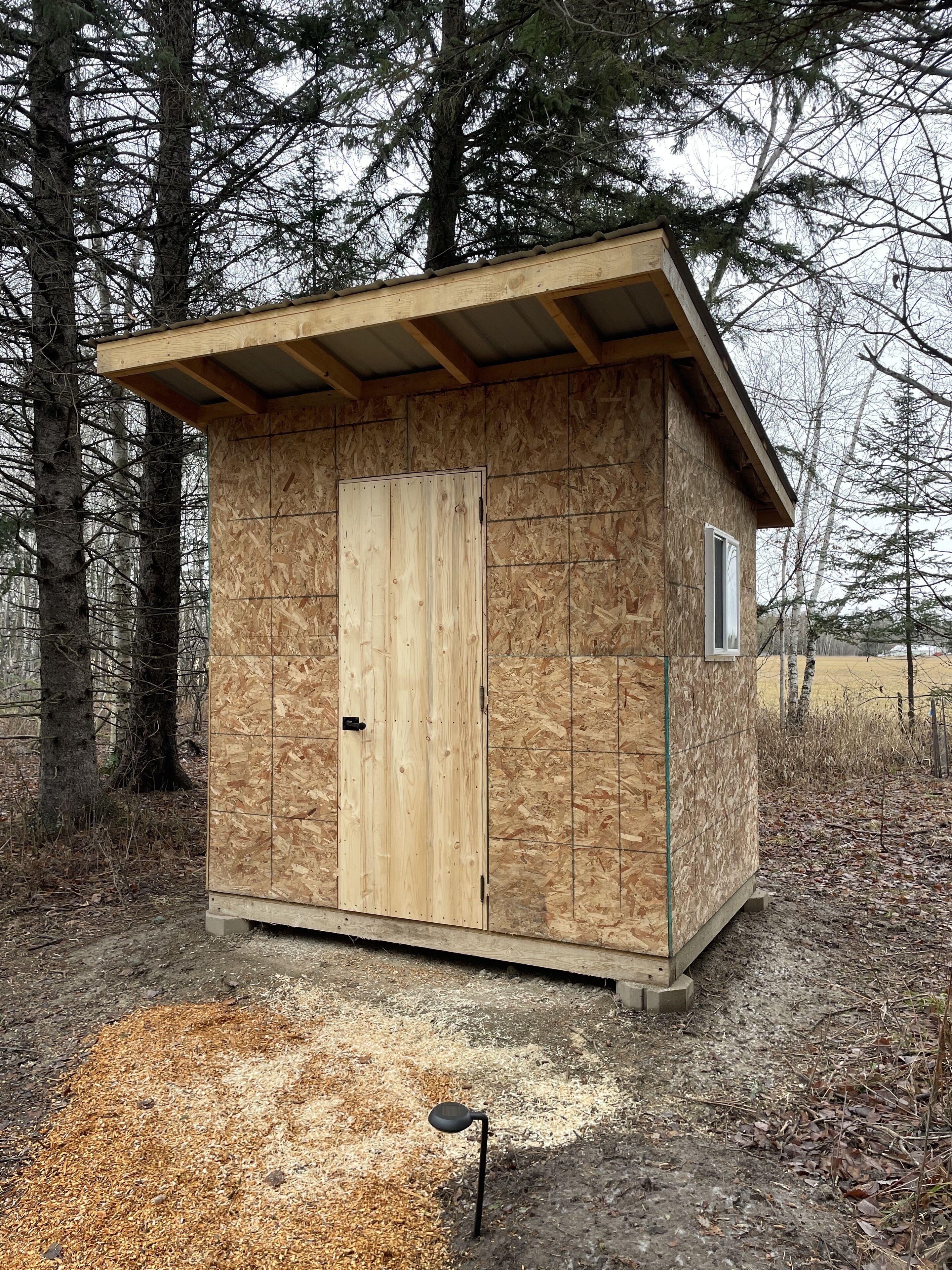 Una pequeña cabaña de madera con una puerta y una ventana, rodeada de árboles.