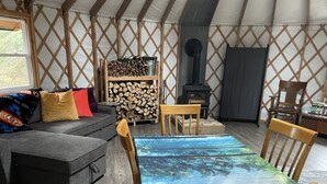 L’intérieur chaleureux d’une yourte, avec un canapé, des chaises en bois, une table ornée d’un motif représentant une forêt, un meuble de rangement pour le bois et un poêle à bois.