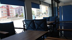 Un área de estar interior moderna con mesas de madera y sillas con cojines azules, amplias ventanas que ofrecen vistas a edificios y a una calle.