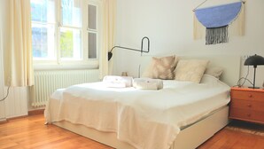 A neatly made bed with a white comforter and pillows, a bedside table with a lamp, and a window with curtains. - Wohnung am Bergpark Wilhelmshöhe (Kassel)