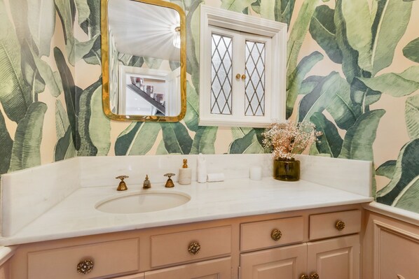 Une salle de bain avec un lavabo blanc, un meuble-lavabo et un miroir avec un cadre doré. On y trouve un meuble avec des portes en verre et un vase rempli de fleurs posé sur le comptoir.