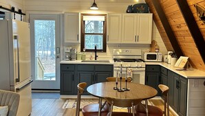 A modern kitchen with wooden beams, a dining area with a round table and stools, and a view of the outdoors through a window. - MCM A-frame: HOT TUB; Lake; Fall Colors; Fireplace; Nature; Cozy; Kayaks (Johannesburg)