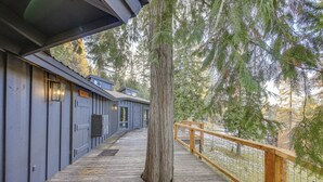 A wooden deck with a railing, a large tree, and a building with a blue facade. - Bonners Ferry Cabin w/ Wraparound Deck & Views! (Bonners Ferry)